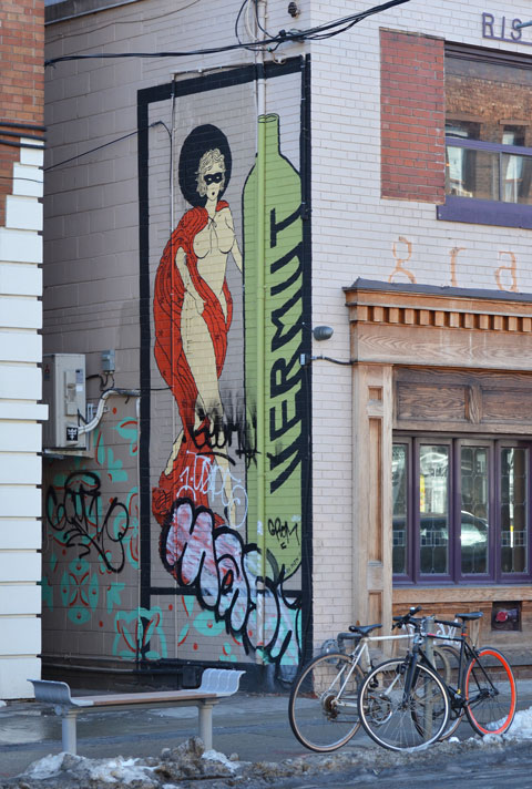 a tall green bottle painted on the wall with the word vermut written vertically. Beside the bottle is painted a naked woman draped in red with breasts showing. 