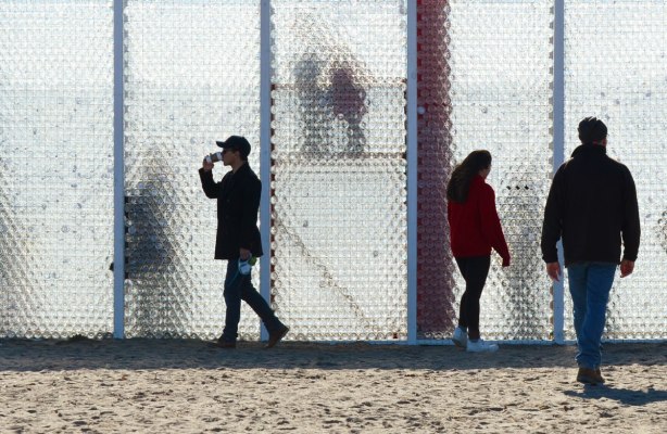 sunlight shines through a wall of plastic bottles, some people walking in front of it. Collective Memory installation at Winter Stations 2017 on Toronto's waterfront. 