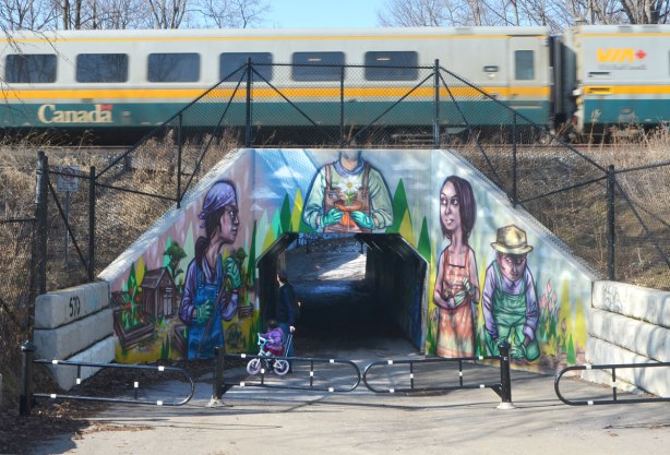a mural by elicser on a railway underpass by the woodfield community rail garden that covers all the concrete of the opening. a VIA train passes overhead as a father and daughter stop to watch 