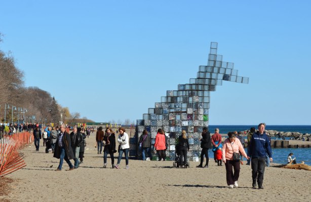 people looking at an art installation on the beach made of wire cage cubes stacked on top of each other. The ones on the bottom are filled with empty plastic bottles of different colours and shapes. The upper cages are empty and they are joined together to look like the head of a creature. 