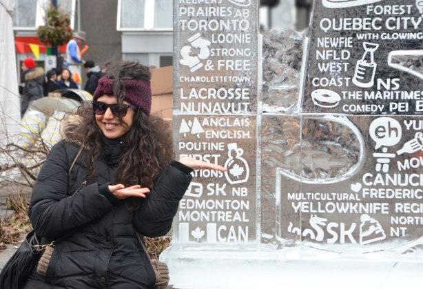 a young woman in black coat and brown tuque stands beside a block of ice that has the names of some of Canada's cities carved into it. 