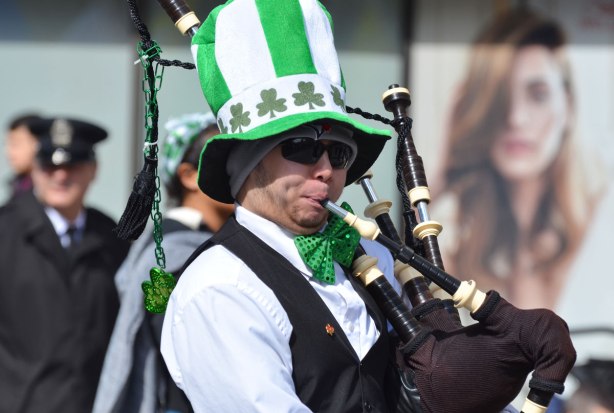 a man playing the bagpipes in a St. Patricks day parade. he's wearing an oversized green and white striped hat with a band of shamrocks on it. Also wearing a big green bowtie