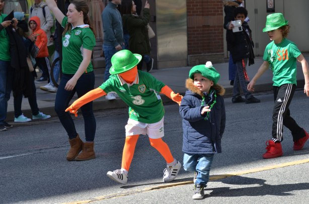 boys running a parade. one is wearing an orange body suit that also covers his face, also green tshirt and white shorts and green bowler. 