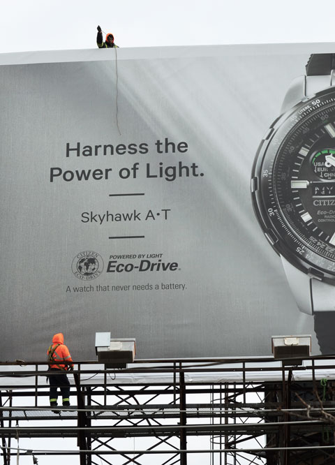 two men are changing the ad on a very large billboard. One man is below and the other is above and he is passing a long rope to man below. 