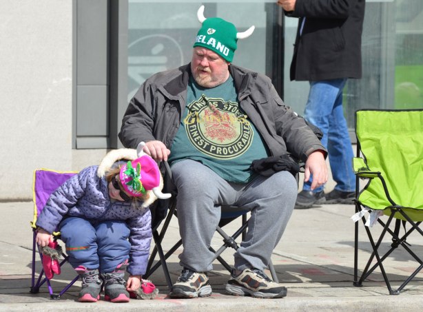 a father and daughter have seats on the sidewalk as they watch a parade go by