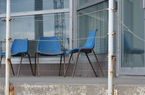 three old blue plastic chairs with metal rusty legs sit on the concrete porch of a commercial building. Windows behind them. one of the chairs is reflected in the window 