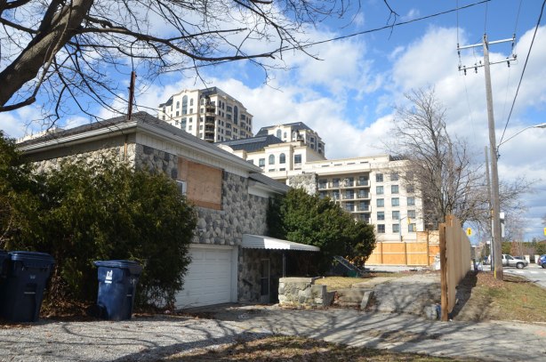 a boarded up house, split level, built in the 1950s, is in the foreground, condos and apartment buildings are behind it 