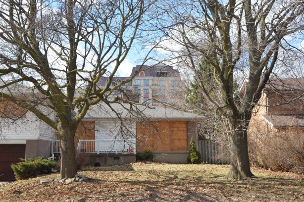 a boarded up house, split level, built in the 1950s, is in the foreground, condos and apartment buildings are behind it 
