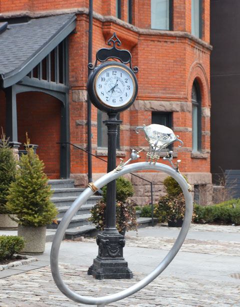 a very large fake diamond ring, single stone, sculpture size, about 3 feet in diameter, stands in front of an old fashioned clock in front of some stores 
