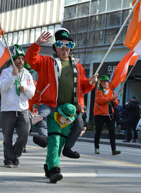 st. patricks day parade, man wearing a costume that makes it look like a large leprechaun is grabbing him around the knees