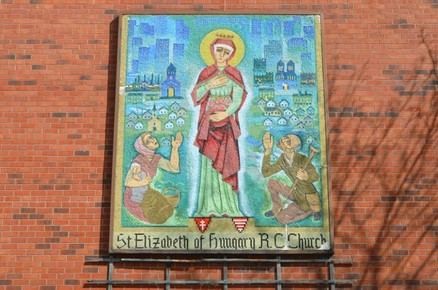 mosaic on the exterior brick wall of St. Elizabeth of Hungary RC Church showing St. Elizabeth and two people kneeling beside her. 