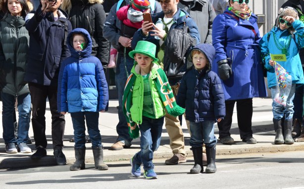 three boys among a crowd of onlookers at a parade. The boy in the middle is wearing green and dancing as he watches 
