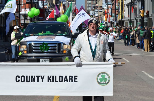 a man holds one end of a white banner that says County Kildare, St. Patricks Day parade 