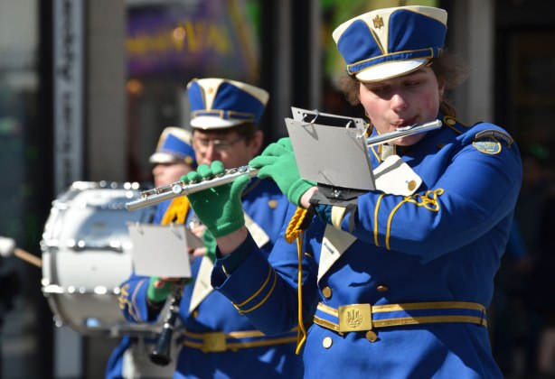 a flute player in a blue uniform plays and walks in a parade. Other musicians in the background