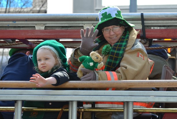 a middle aged woman in a fireman's jacket sits on a float in a parade (firetruck?) whe is holding a small teddy bear with a green sweater and a young girl is with her