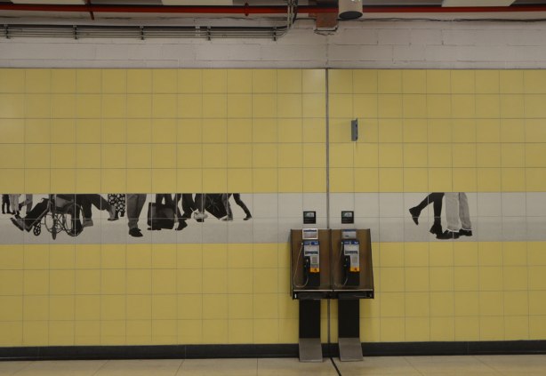 photos of peoples legs and feet in black and white on tiles on a yellow tiled subway station wall, over and beside two Bell pay phones, artwork by Sylvie Belanger