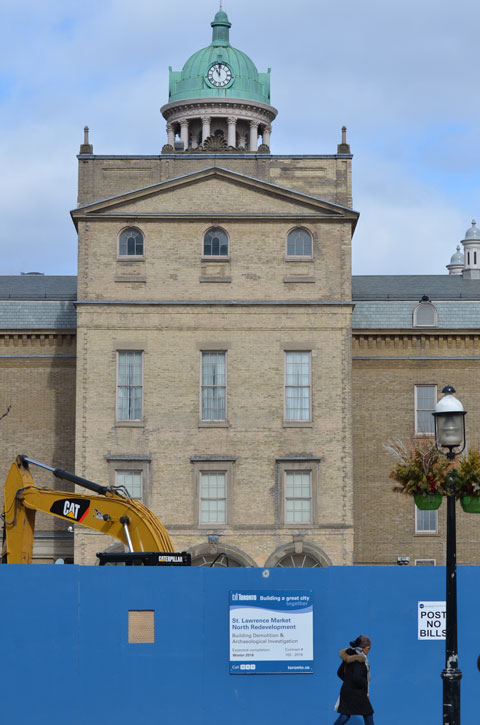 the rear of the old St. Lawrence Hall building, with a bright blue wood hoarding fence in front of it. a woman is walking past 