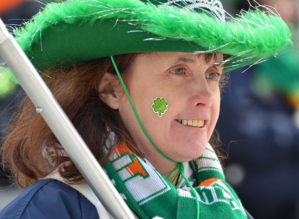 a woman in a green cowboy hat with a green fringe, a green shamrock sticker on her cheek and wearing a green, orange and white scarf