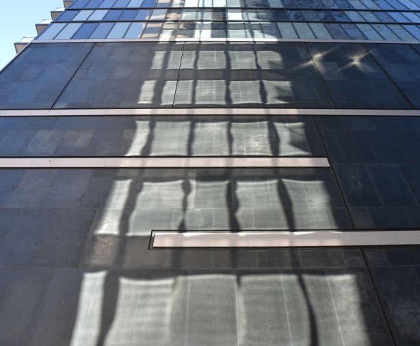 looking up a tall building that is black on the exterior and has light reflected from a glass building beside it.