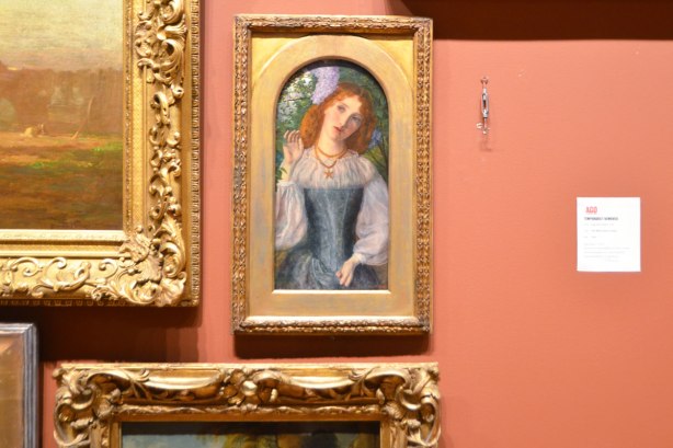 small painting of a red headed woman torso, amrs, and head, in a gold frame on a wall with lots of other paintings, except on e is missing and there is a white sign saying why its missing