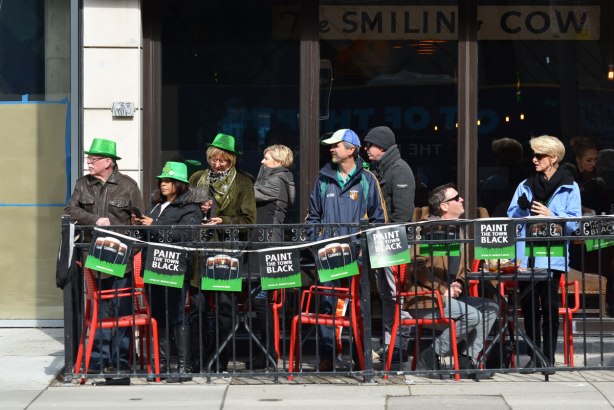 people standing and sitting on a small outdoor patio, on the sidewalk, watching the St. Patricks day parade go past. The bar is the Smiling Cow.