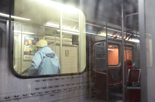 reflections of a woman in a red jacket sitting on the subway, reflected in the window beside a woman who is standing on the platform 