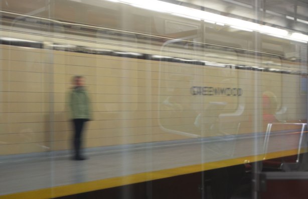 blurry person standing on the platform at Greenwood station, with reflections of people sitting on the subway