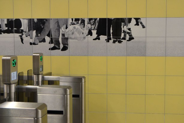 photos of peoples legs and feet in black and white on tiles on a yellow tiled subway station wall, over the metal turnstiles for entry into the station, artwork by Sylvie Belanger