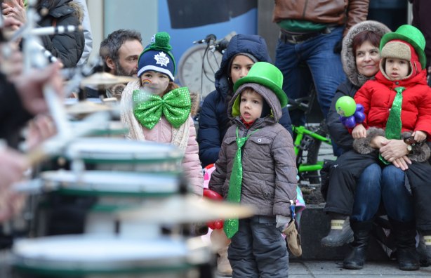 adults and kids sitting on the sidewalk watch drummers go past in a parade. girl with big green bowtie and boy with long green tie and green bowler hat
