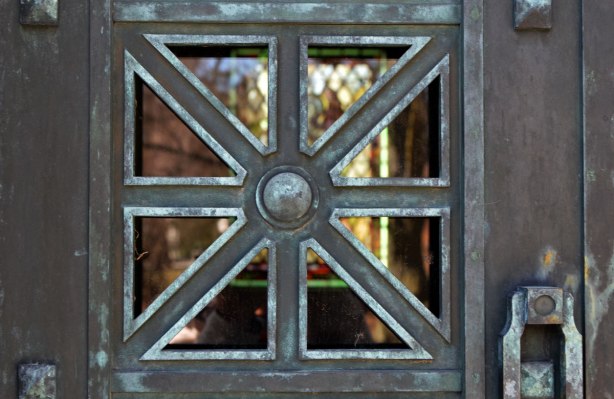 close up of a pane in a window with a metal window frame, square with lines dividing the pane into 8 triangles, stained glass window in the background. Looking into a vault at a cemetery