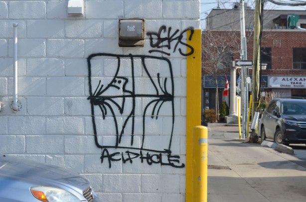 the front of a grey car parked in front of a white wall in an alley where there is a line drawing graffiti of a man peering from behind the curtains of a window.