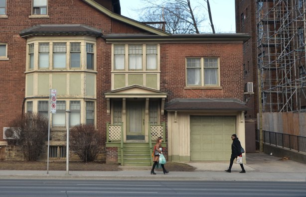 two women walk past a brick house with green wood features, porch, windows, garage door. 