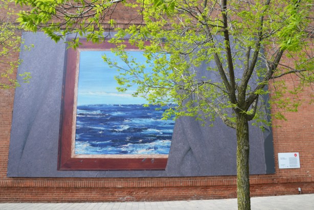 a large art installation on the south exterior wall of the Power Plant contemporary art gallery, with a small tree in front of it. 