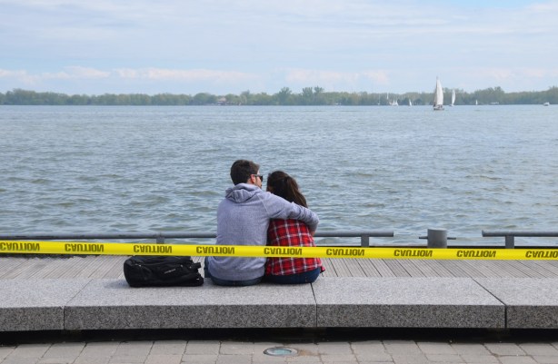 a young couple sits by the waterfront, on a stone bench. He has his arm around her. There is yellow caution tape behind them because the water level in Lake Ontario is high. 