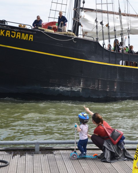 a mother crouches down beside a young child who is wearing a helmet and is on a scooter, the mother is waving at the Kajama as it docks, the Kajama is a boat with sails that gives tourists rides on Lake Ontario