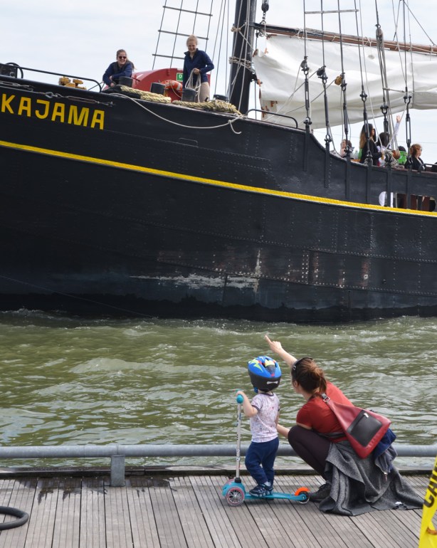 a mother crouches down beside a young child who is wearing a helmet and is on a scooter, the mother is waving at the Kajama as it docks, the Kajama is a boat with sails that gives tourists rides on Lake Ontario