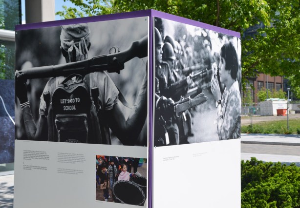 2 sides, taken from the corner, of a box like structure, with black and white photographs on the two sides, one of the back of a man with a rifle across his shoulders and a backpack that says Lets go to school. The other photo is a woman standing up to a line of soldiers with bayonets. 