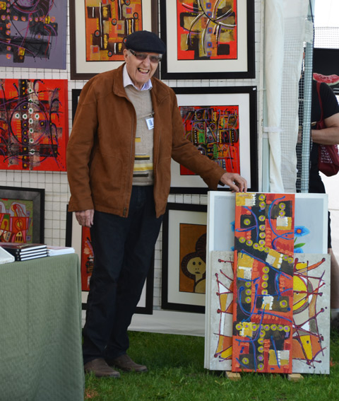 a man stands holding up a painting (bottom of painting is resting on the ground), more painting displayed on the wall behind him, at the outdoor Riverdale Artwalk art show and sale 