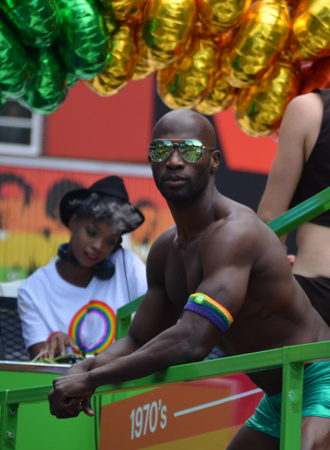 a black man wearing just green tight shorts and green sunglasses is standing on the TD float, a woman is in the background (she is looking after the music). 