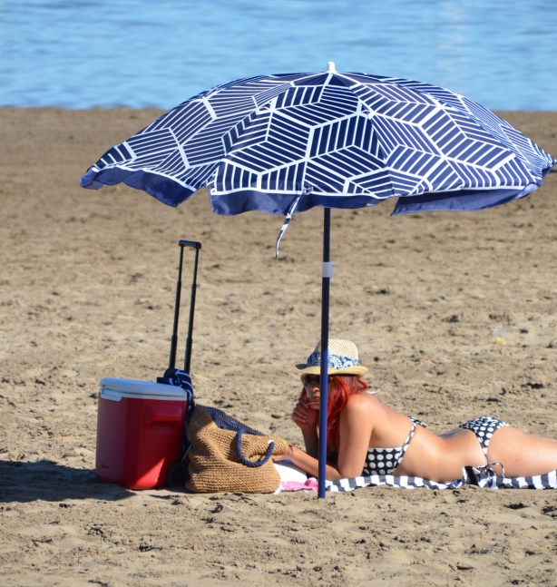 a woman with red hair lies under a blue and white umbrella on the beach by Lake Ontario