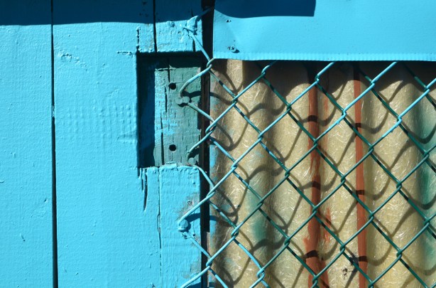 chain link fence nailed to a bright blue wood fence, corrugated plastic behind the chain link 
