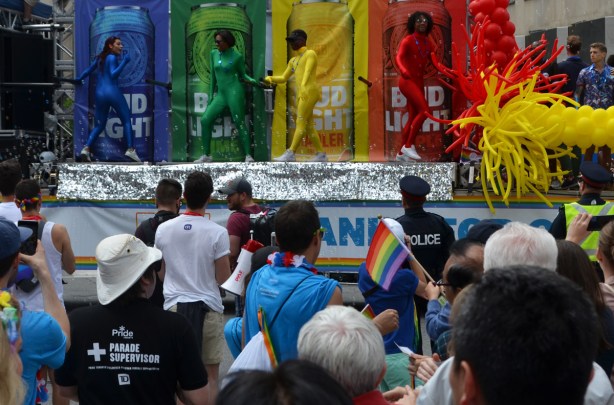 bud light float at pride, picture of 4 large cans - a blue, green, red, and yellow can with a person in front of each in a tight body suit the same colour as the can. crowd in front of the float.