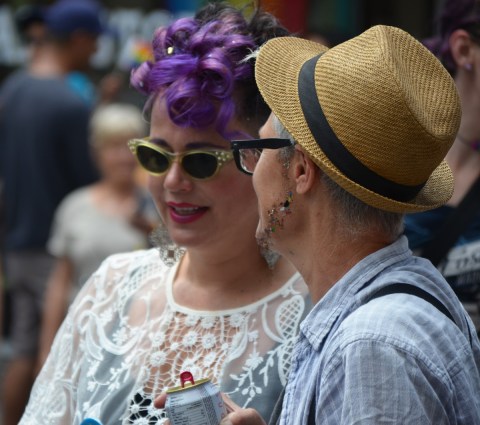 a couple - one is a purple wig and sunglasses and the other in a straw hat with sparkles glued to her face in the shape of a thin beard