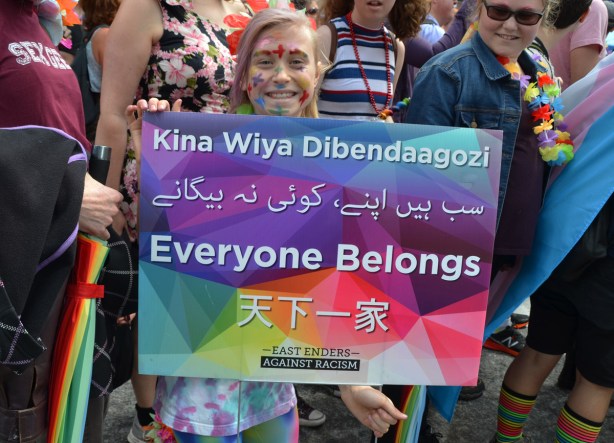 a girl holds up a sign from the East Enders Everyone belongs, that has the expression everyone belongs, written in an indigenous language as well as arabic and chinese (or other Asian language)