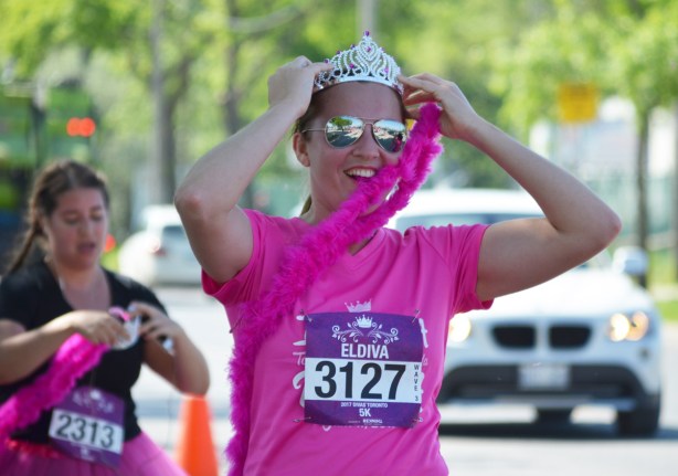 a young woman in a pink T-shirt, runner 3127, Eldiva, puts on her tiara and pink boa as she nears the end of the Run Like a Diva 