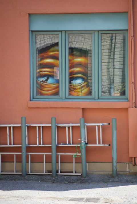 an orange, or salmon, coloured wall with a window. in the window is a reflection of a pair of eyes from a large street art mural. under the window are two ladders lying horizontal. 