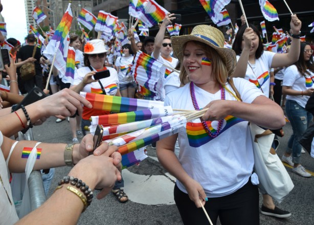 a woman in a white t shirt with rainbow on the front, and a straw hat is passing out rainbow flags to the crowd. others in the parade behind her are doing the same, others are waving the flags.
