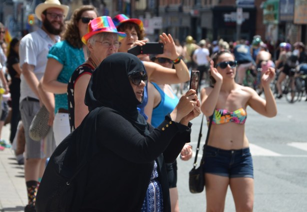 a muslim woman in a black head scarf takes pictures on her phone at a dyke march. a woman in a bikini top is clapping as she walks toward the camera