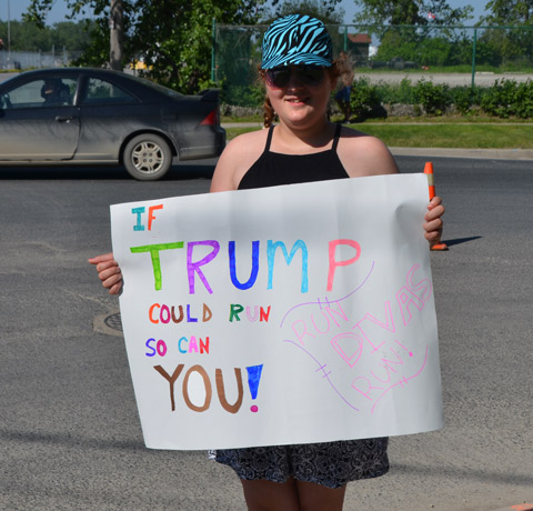 a young woman, a spectator at a 5km run, holds a sign that says If Trump can run, so can you