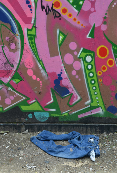 a pair of blue jeans are lying on the ground at the foot of a wall that has pink and green street art on it. 